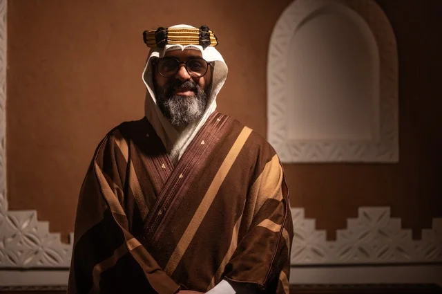 ايماءات الوقار والسرور ، رجل عربي سعودي مسن ينظر إلى الكاميرا يرتدي الزي العربي التقليدي يقف داخل غرفة تراثية بديكورات جدران مزخرفة، يرتدي عباءة واسعة وعقالًا على رأسه، ويبدو بلحية بيضاء، الحكمة والتجربة الطويلة في الحياة، الأصالة والحنين للأيام القديمة