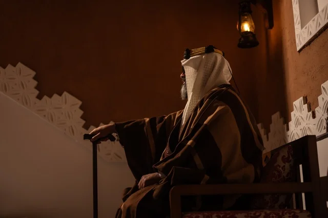 الحكمة والتجربة الطويلة في الحياة، صورة جانبية لرجل عربي سعودي مسن يرتدي الزي العربي التقليدي يجلس على كرسي خشبي داخل غرفة تراثية بديكورات جدران مزخرفة، يرتدي عباءة واسعة وعقالًا على رأسه، ويبدو بلحية بيضاء، بيده عكاز يستند إليها .