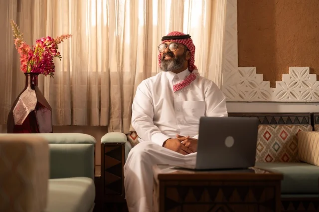 The atmosphere of comfort and remote work, the meaning of authenticity and modernity, a Saudi Arabian Gulf man sitting inside a traditional council adorned with wall decorations and traditional ornaments, wearing a white thobe with a red shemagh and agal, contemplating the sunlight, with an open laptop in front of him.
