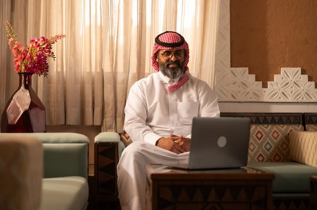 A traditional Arab environment enhanced with modern touches, embodying the meaning of authenticity and modernity. A Saudi Gulf Arab man sits inside a heritage council adorned with wall decorations and traditional ornaments, in a setting of comfort and remote work. He is wearing a white thobe with a red shemagh and agal, with an open laptop in front of him.
