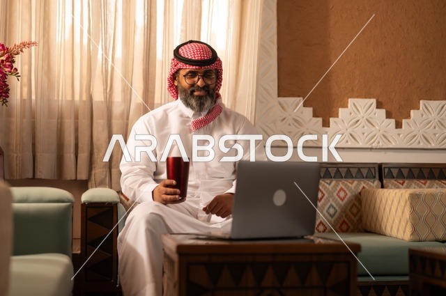 أجواء الراحة والعمل عن بعد، معنى الأصالة والحداثة، رجل عربي خليجي أنيق يجلس داخل مجلس تراثي مزين بديكورات جدارية وزخارف تقليدية، يرتدي الثوب الأبيض مع الشماغ الأحمر والعقال يمسك بيده كوباً معدنياَ ، أمامه جهاز حاسوب محمول مفتوح،