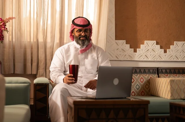 أجواء الراحة والعمل عن بعد، معنى الأصالة والحداثة، رجل عربي خليجي أنيق يجلس داخل مجلس تراثي مزين بديكورات جدارية وزخارف تقليدية، يرتدي الثوب الأبيض مع الشماغ الأحمر والعقال يمسك بيده كوباً معدنياَ ، أمامه جهاز حاسوب محمول مفتوح، أجواء الراحة والعمل عن بعد، معنى الأصالة والحداثة، رجل عربي خليجي أنيق يجلس داخل مجلس تراثي مزين بديكورات جدارية وزخارف تقليدية، يرتدي الثوب الأبيض مع الشماغ الأحمر والعقال يمسك بيده كوباً معدنياَ ، أمامه جهاز حاسوب محمول مفتوح،