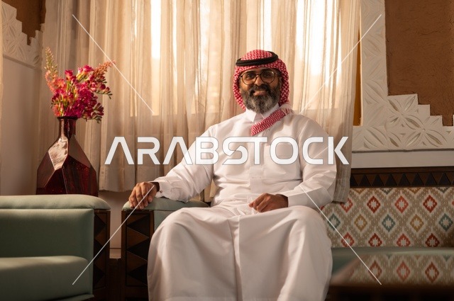 ايماءات الثقة والسرور، الأرائك المزخرفة والمزهريات التي تحمل وروداً ملونة، رجل عربي خليجي سعودي أنيق يجلس داخل مجلس تراثي مزين بديكورات جدارية وزخارف تقليدية، أجواء الراحة والعمل عن بعد، يرتدي الثوب الأبيض مع الشماغ الأحمر والعقال