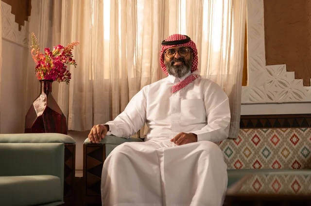 ايماءات الثقة والسرور، الأرائك المزخرفة والمزهريات التي تحمل وروداً ملونة، رجل عربي خليجي سعودي أنيق يجلس داخل مجلس تراثي مزين بديكورات جدارية وزخارف تقليدية، أجواء الراحة والعمل عن بعد، يرتدي الثوب الأبيض مع الشماغ الأحمر والعقال