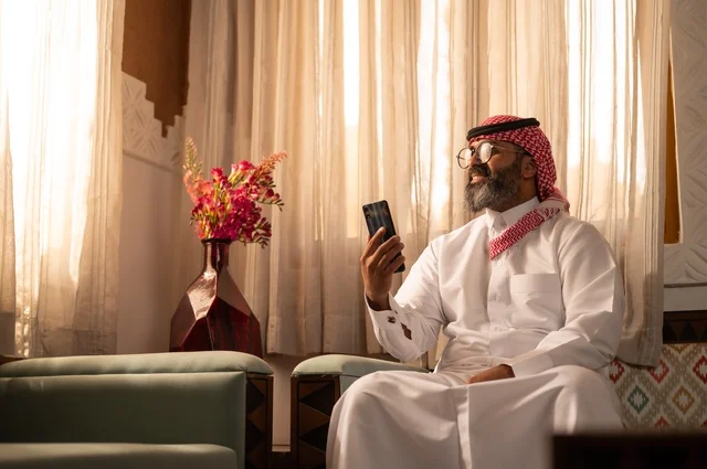 الأرائك المزخرفة والمزهريات التي تحمل وروداً ملونة، استخدام التكنولوجيا في الحياة اليومية، رجل عربي خليجي سعودي أنيق يجلس داخل مجلس تراثي مزين بديكورات جدارية وزخارف تقليدية، أجواء الراحة والعمل عن بعد، يرتدي الثوب الأبيض مع الشماغ الأحمر والعقال