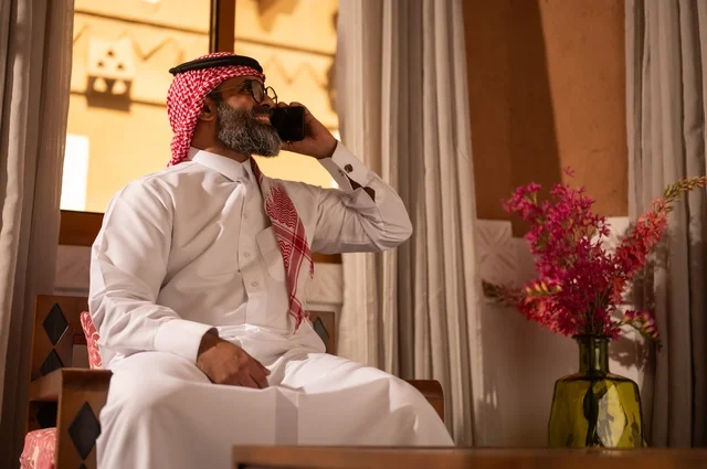 Making a phone call, a traditional Arab environment enhanced with modern touches, the meaning of authenticity and modernity, a Saudi Gulf Arab man sitting inside a heritage council decorated with wall decorations and traditional ornaments, an atmosphere of comfort and remote work, wearing a white thobe with a red shemagh and agal.