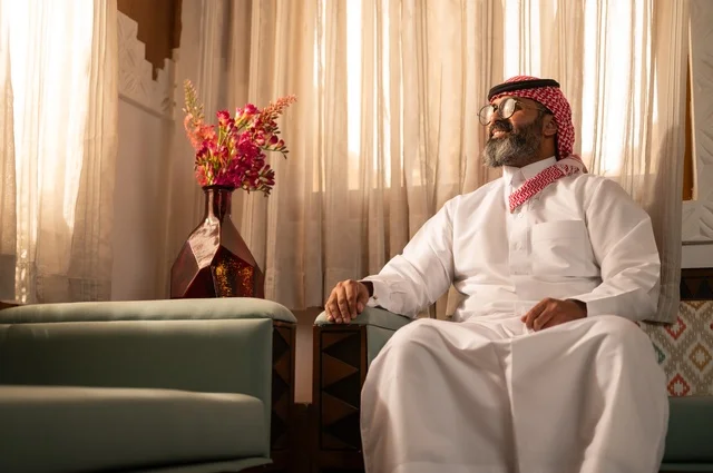 A warm atmosphere filled with life and cultural identity, decorated sofas and vases holding colorful flowers, a Saudi Arabian Gulf man sitting inside a traditional council adorned with wall decorations and traditional motifs, an ambiance of comfort and remote work, wearing a white thobe with a red shemagh and agal.