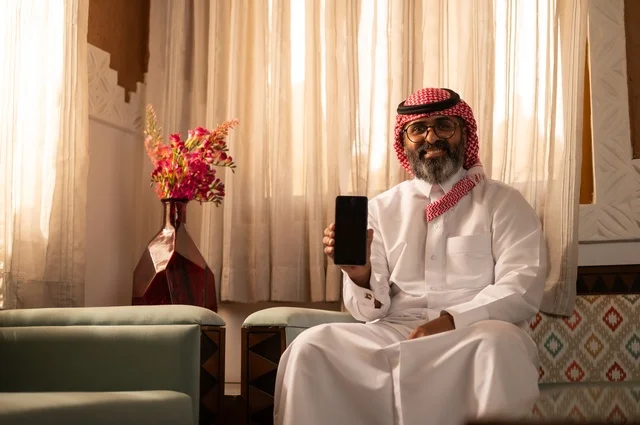 عرض خلفية سوداء على الهاتف المحمول، بيئة عربية تقليدية مزودة بلمسات عصري، معنى الأصالة والحداثة، رجل عربي خليجي سعودي يجلس داخل مجلس تراثي مزين بديكورات جدارية وزخارف تقليدية، أجواء الراحة والعمل عن بعد، يرتدي الثوب الأبيض مع الشماغ الأحمر والعقال