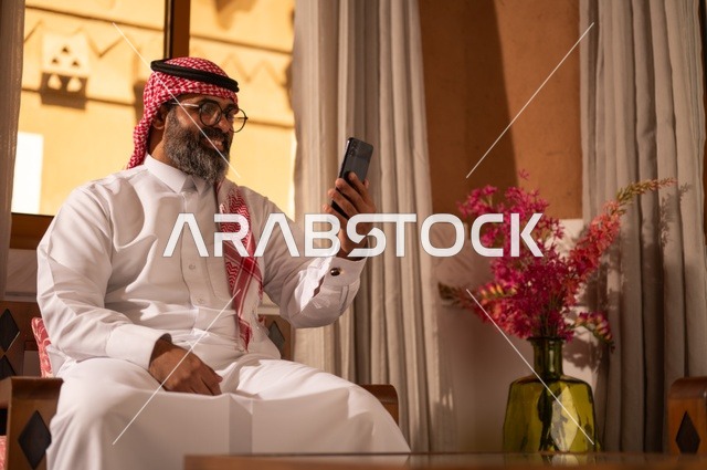 استخدام التكنولوجيا في الحياة اليومية، بيئة عربية تقليدية مزودة بلمسات عصري، معنى الأصالة والحداثة، رجل عربي خليجي سعودي  يجلس داخل مجلس تراثي مزين بديكورات جدارية وزخارف تقليدية، أجواء الراحة والعمل عن بعد، يرتدي الثوب الأبيض مع الشماغ الأحمر والعقال