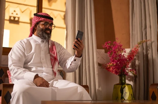 The use of technology in daily life, a traditional Arab environment enhanced with modern touches, the meaning of authenticity and modernity, a stylish Gulf Arab man sitting inside a heritage council adorned with wall decorations and traditional ornaments, an atmosphere of comfort and remote work, wearing a white thobe with a red ghutrah and agal.