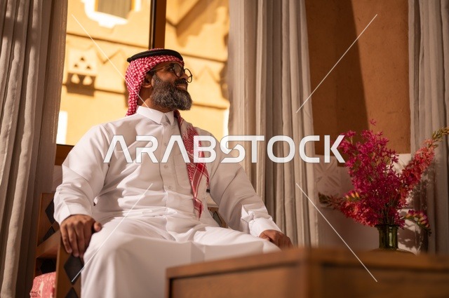رجل عربي خليجي سعودي يجلس داخل مجلس تراثي مزين بديكورات جدارية وزخارف تقليدية، تعابير الراحة والاسترخاء، يرتدي الثوب الأبيض مع الشماغ الأحمر والعقال الأرائك المزخرفة والمزهريات التي تحمل وروداً ملونة، الأناقة والجمال