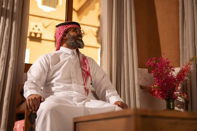 رجل عربي خليجي سعودي يجلس داخل مجلس تراثي مزين بديكورات جدارية وزخارف تقليدية، تعابير الراحة والاسترخاء، يرتدي الثوب الأبيض مع الشماغ الأحمر والعقال الأرائك المزخرفة والمزهريات التي تحمل وروداً ملونة، الأناقة والجمال