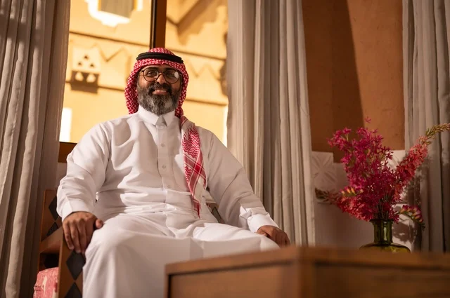 تعابير وجه ابتسامة وسرور، رجل عربي خليجي سعودي يجلس داخل مجلس تراثي مزين بديكورات جدارية وزخارف تقليدية، تعابير الراحة والاسترخاء، يرتدي الثوب الأبيض مع الشماغ الأحمر والعقال الأرائك المزخرفة والمزهريات التي تحمل وروداً ملونة، الأناقة والجمال