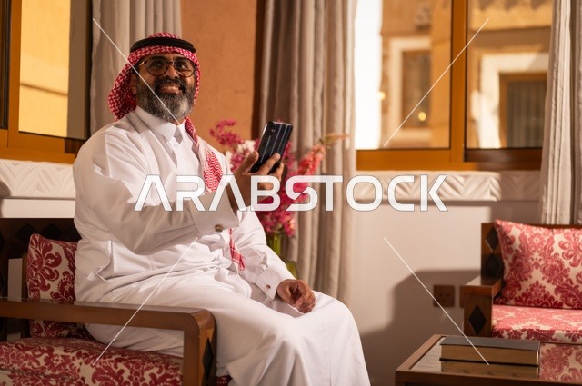 استخدام التكنولوجيا في الحياة اليومية، بيئة عربية تقليدية مزودة بلمسات عصري، معنى الأصالة والحداثة، رجل عربي خليجي سعودي أنيق يجلس داخل مجلس تراثي مزين بديكورات جدارية وزخارف تقليدية، أجواء الراحة والعمل عن بعد، يرتدي الثوب الأبيض مع الشماغ الأحمر والعقال