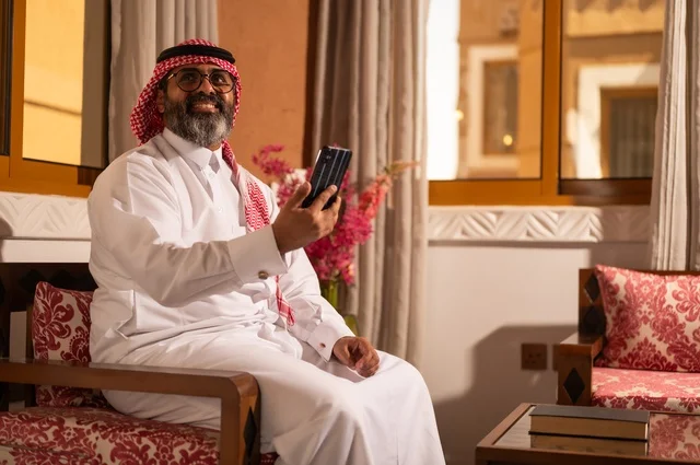 استخدام التكنولوجيا في الحياة اليومية، بيئة عربية تقليدية مزودة بلمسات عصري، معنى الأصالة والحداثة، رجل عربي خليجي سعودي أنيق يجلس داخل مجلس تراثي مزين بديكورات جدارية وزخارف تقليدية، أجواء الراحة والعمل عن بعد، يرتدي الثوب الأبيض مع الشماغ الأحمر والعقال