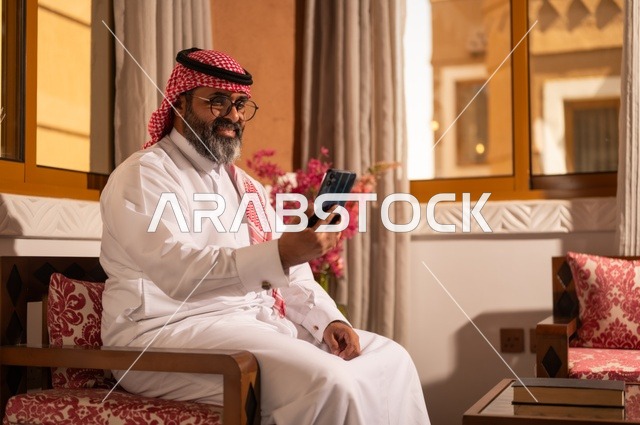 الأرائك المزخرفة والمزهريات التي تحمل وروداً ملونة، استخدام التكنولوجيا في الحياة اليومية، رجل عربي خليجي سعودي أنيق يجلس داخل مجلس تراثي مزين بديكورات جدارية وزخارف تقليدية، أجواء الراحة والعمل عن بعد، يرتدي الثوب الأبيض مع الشماغ الأحمر والعقال
