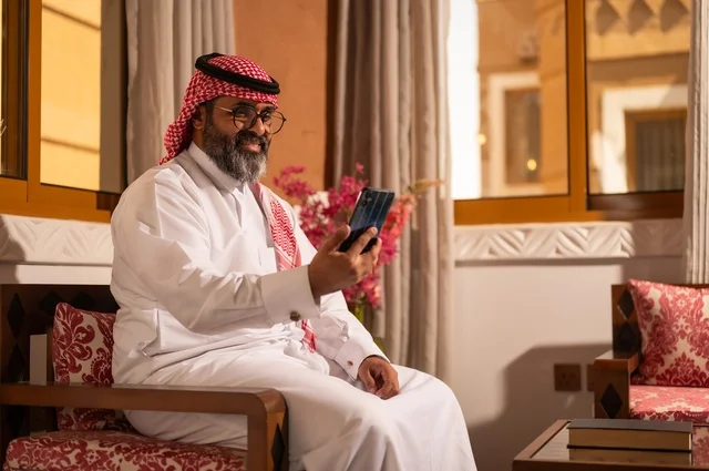 الأرائك المزخرفة والمزهريات التي تحمل وروداً ملونة، استخدام التكنولوجيا في الحياة اليومية، رجل عربي خليجي سعودي أنيق يجلس داخل مجلس تراثي مزين بديكورات جدارية وزخارف تقليدية، أجواء الراحة والعمل عن بعد، يرتدي الثوب الأبيض مع الشماغ الأحمر والعقال
