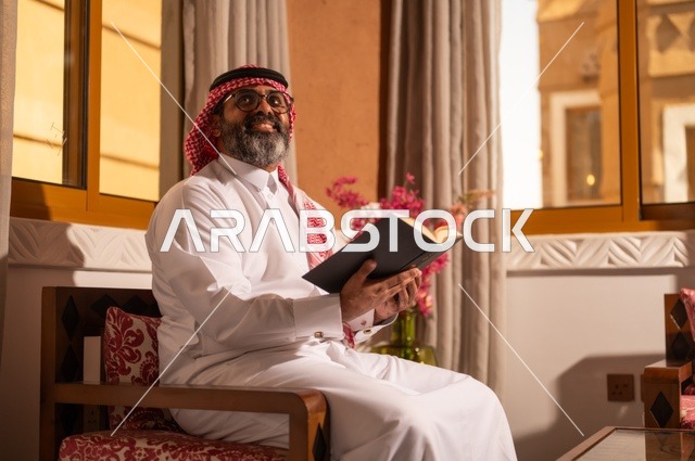 صورة جانبية لرجل عربي خليجي يجلس داخل مجلس تراثي بديكورات عربية وزخارف تقليدية يمسك بيده كتاباً يقرأ فيه، النظر إلى الكاميرا بتعابير وجه مبهجة، قيمة القراءة والاطلاع، الثقافة وحب المعرفة والارتباط بالعلم، يرتدي الثوب الأبيض والشماغ الأحمر والعقال