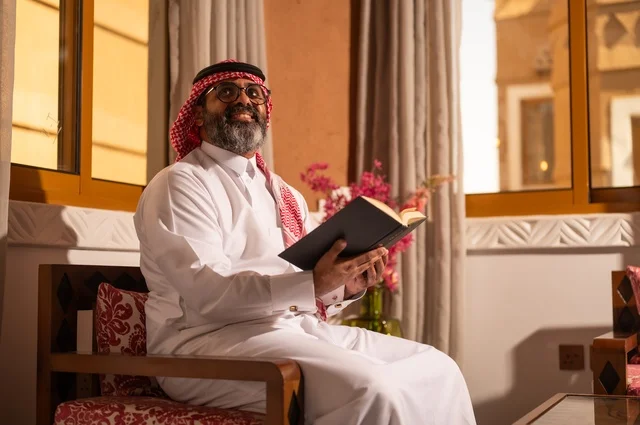 A side image of an Arab Gulf man sitting inside a traditional council with Arab decorations and traditional ornaments, holding a book he is reading, looking at the camera with a joyful expression on his face, reflecting the value of reading and knowledge, culture, love for knowledge, and connection to science, wearing a white thobe, a red ghutrah, and an agal.