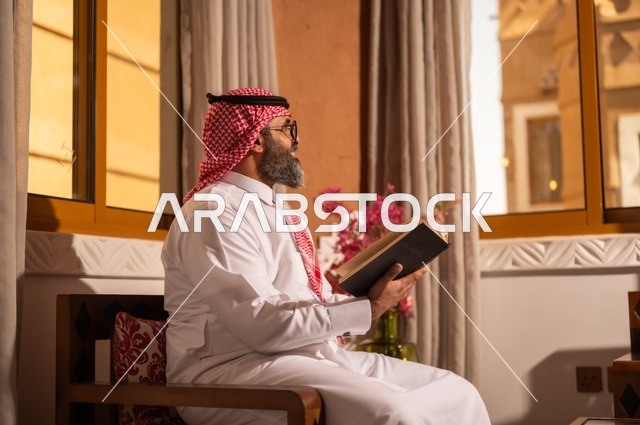 قيمة القراءة والاطلاع، الثقافة وحب المعرفة والارتباط بالعلم، رجل عربي خليجي يجلس داخل مجلس تراثي بديكورات عربية وزخارف تقليدية، يرتدي الثوب الأبيض والشماغ الأحمر والعقال، ويضع نظارات طبية، يمسك بيده كتاباً يقرأ فيه باهتمام، تفاصيل الأرائك المزخرفة