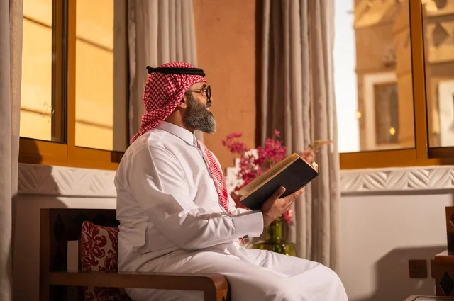 قيمة القراءة والاطلاع، الثقافة وحب المعرفة والارتباط بالعلم، رجل عربي خليجي يجلس داخل مجلس تراثي بديكورات عربية وزخارف تقليدية، يرتدي الثوب الأبيض والشماغ الأحمر والعقال، ويضع نظارات طبية، يمسك بيده كتاباً يقرأ فيه باهتمام، تفاصيل الأرائك المزخرفة