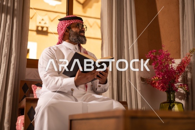 رجل عربي خليجي يجلس داخل مجلس تراثي بديكورات عربية وزخارف تقليدية يمسك بيده كتاباً يقرأ فيه، يضع نظارة طبية، قيمة القراءة والاطلاع، الثقافة وحب المعرفة والارتباط بالعلم، يرتدي الثوب الأبيض والشماغ الأحمر والعقال