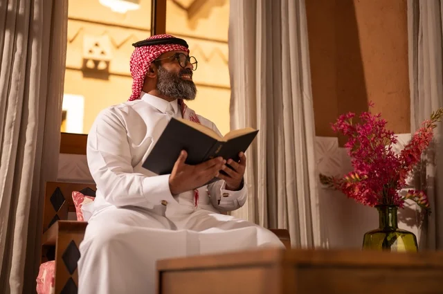 A Gulf Arab man is sitting inside a traditional council with Arabic decorations and traditional motifs, holding a book in his hand that he is reading. He is wearing reading glasses, emphasizing the value of reading and knowledge, culture, and a love for learning and connection to science. He is dressed in a white thobe, a red ghutrah, and an agal.
