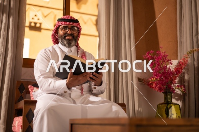 النظر إلى الكاميرا بتعابير وجه مبهجة، قيمة القراءة والاطلاع، الثقافة وحب المعرفة والارتباط بالعلم، رجل عربي خليجي يجلس داخل مجلس تراثي بديكورات عربية وزخارف تقليدية، يرتدي الثوب الأبيض والشماغ الأحمر والعقال، ويضع نظارات طبية، يمسك بيده كتاباً يقرأ فيه