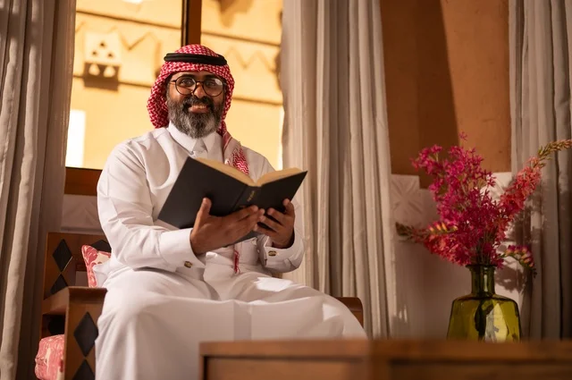 النظر إلى الكاميرا بتعابير وجه مبهجة، قيمة القراءة والاطلاع، الثقافة وحب المعرفة والارتباط بالعلم، رجل عربي خليجي يجلس داخل مجلس تراثي بديكورات عربية وزخارف تقليدية، يرتدي الثوب الأبيض والشماغ الأحمر والعقال، ويضع نظارات طبية، يمسك بيده كتاباً يقرأ فيه