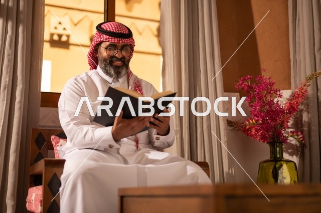 Culture, love of knowledge, and connection to science. An Arab Gulf man sits inside a traditional council with Arab decorations and traditional ornaments. He wears a white thobe, a red ghutrah, and an agal, and he has on reading glasses. He holds a book in his hand, reading it with interest, surrounded by details of the ornate sofas, emphasizing the value of reading and exploration.