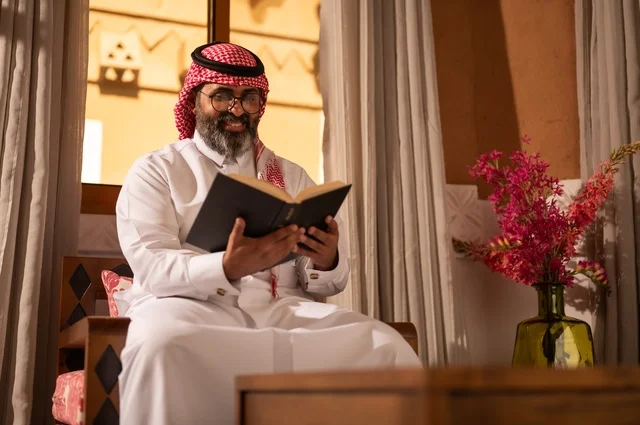 الثقافة وحب المعرفة والارتباط بالعلم، رجل عربي خليجي يجلس داخل مجلس تراثي بديكورات عربية وزخارف تقليدية، يرتدي الثوب الأبيض والشماغ الأحمر والعقال، ويضع نظارات طبية، يمسك بيده كتاباً يقرأ فيه باهتمام، تفاصيل الأرائك المزخرفة ، قيمة القراءة والاطلاع