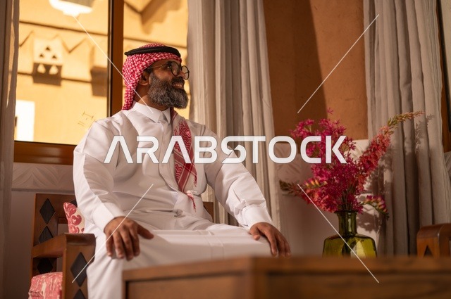 تعابير الراحة والاسترخاء، الأرائك المزخرفة والمزهريات التي تحمل وروداً ملونة، رجل عربي خليجي سعودي أنيق يجلس داخل مجلس تراثي مزين بديكورات جدارية وزخارف تقليدية، أجواء الراحة والعمل عن بعد، يرتدي الثوب الأبيض مع الشماغ الأحمر والعقال