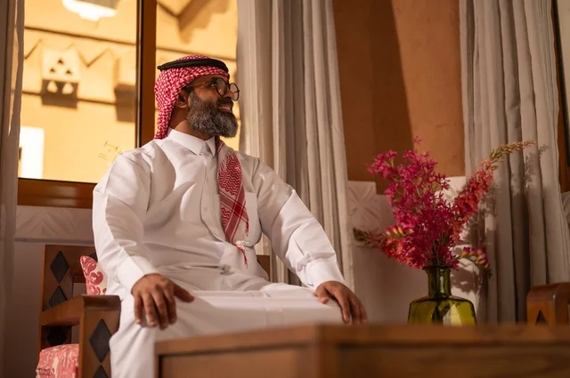 Expressions of comfort and relaxation, ornate sofas and vases carrying colorful flowers, a stylish Arab Gulf Saudi man sitting inside a traditional council adorned with wall decorations and traditional motifs, an atmosphere of comfort and remote work, wearing a white thobe with a red shemagh and agal.
