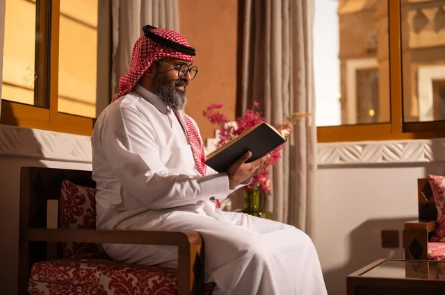 قيمة القراءة والاطلاع، الثقافة وحب المعرفة والارتباط بالعلم، رجل عربي خليجي يجلس داخل مجلس تراثي بديكورات عربية وزخارف تقليدية، يرتدي الثوب الأبيض والشماغ الأحمر والعقال، ويضع نظارات طبية، يمسك بيده كتاباً يقرأ فيه باهتمام، تفاصيل الأرائك المزخرفة