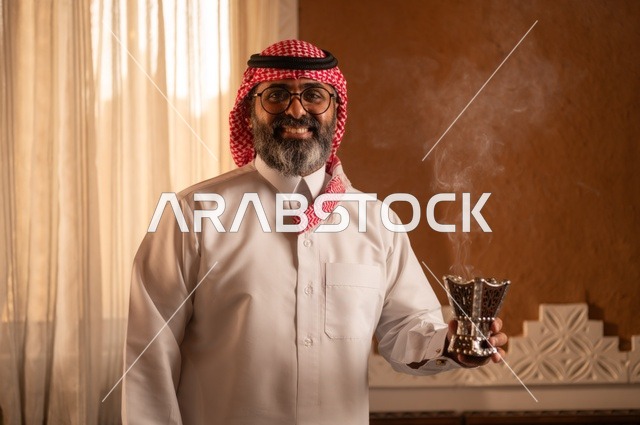 صورة مقربة لرجل عربي خليجي يقف داخل مجلس تراثي بديكورات عربية وزخارف تقليدية يمسك بيده مبخرة عربية، يضع نظارة طبية، العود والروائح العربية، توازناً جميلاً بين الأصالة العربية والجمال، يرتدي الثوب الأبيض والشماغ الأحمر والعقال