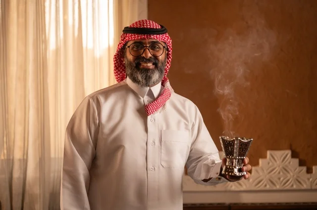 صورة مقربة لرجل عربي خليجي يقف داخل مجلس تراثي بديكورات عربية وزخارف تقليدية يمسك بيده مبخرة عربية، يضع نظارة طبية، العود والروائح العربية، توازناً جميلاً بين الأصالة العربية والجمال، يرتدي الثوب الأبيض والشماغ الأحمر والعقال
