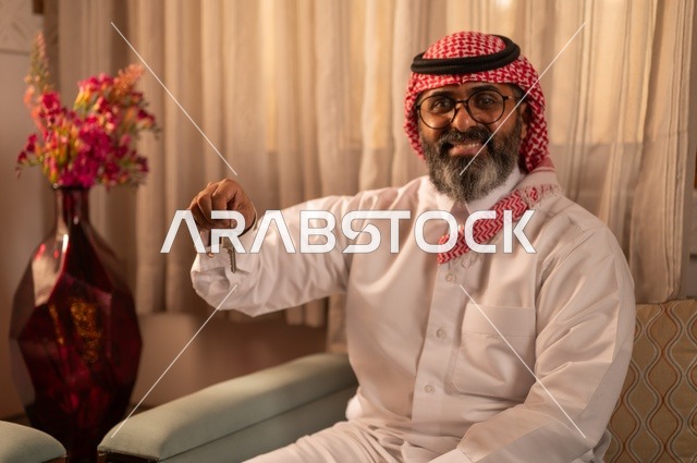 الملكية والأمان والحياة التقليدية، رجل عربي خليجي يجلس داخل مجلس تراثي بديكورات عربية وزخارف تقليدية يمسك بيده مفتاح منزل، يضع نظارة طبية، توازناً جميلاً بين الأصالة العربية والجمال، يرتدي الثوب الأبيض والشماغ الأحمر والعقال