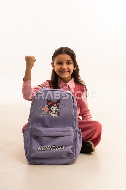 التجهز للذهاب الى اليوم الاول في المدرسة، حب التعلم و التعليم, الاستعداد للعام الدراسي الجديد,  تعابير وإيماءات السعادة و السرور, بورتريه لفتاة عربية خليجية سعودية مبتسمه ترتدي الزي المدرسي تقوم بتوضيب حقيبتها المدرسية، خلفية بيضاء
