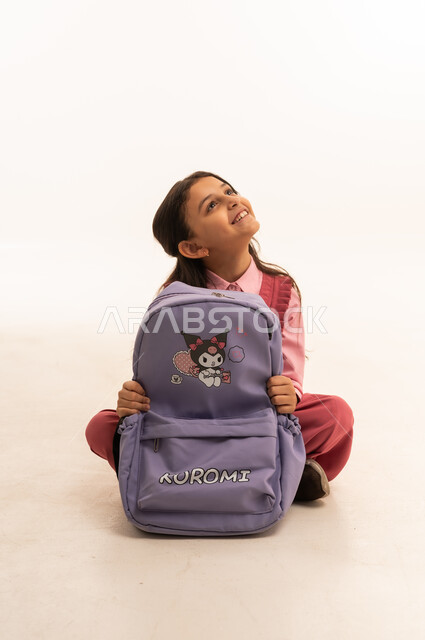 التجهز للذهاب الى اليوم الاول في المدرسة، حب التعلم و التعليم, الاستعداد للعام الدراسي الجديد,  تعابير وإيماءات السعادة و السرور, بورتريه لفتاة عربية خليجية سعودية مبتسمه ترتدي الزي المدرسي تقوم بتوضيب حقيبتها المدرسية، خلفية بيضاء