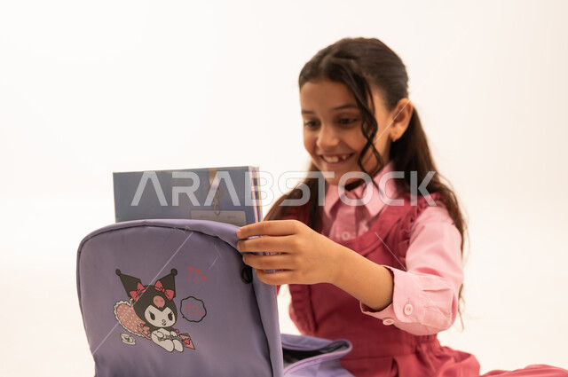  الاستعداد للعام الدراسي الجديد,  تعابير وإيماءات السعادة و السرور, بورتريه لفتاة عربية خليجية سعودية مبتسمه ترتدي الزي المدرسي تقوم بتوضيب حقيبتها المدرسية، التجهز للذهاب الى اليوم الاول في المدرسة، حب التعلم و التعليم, خلفية بيضاء