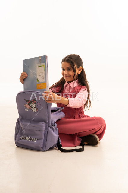  الاستعداد للعام الدراسي الجديد,  تعابير وإيماءات السعادة و السرور, بورتريه لفتاة عربية خليجية سعودية مبتسمه ترتدي الزي المدرسي تقوم بتوضيب حقيبتها المدرسية، التجهز للذهاب الى اليوم الاول في المدرسة، حب التعلم و التعليم, خلفية بيضاء