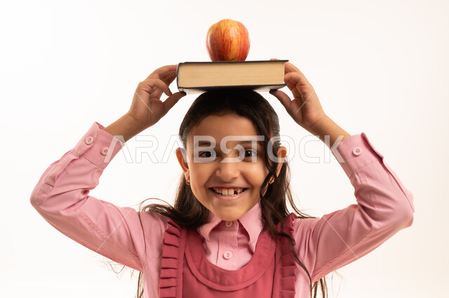 Eating healthy fruits, preparing for the new school year, loving learning and education, looking at the camera with gestures of happiness and pleasure, close-up portrait of a Saudi Arabian Gulf girl wearing a school uniform with school books and a green apple on her head, white background