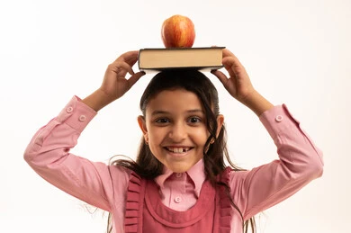 تناول الفواكه الصحية, الاستعداد للعام الدراسي الجديد، حب التعلم و التعليم, النظر إلى الكاميرا بإيماءات السعادة و السرور, بورتريه مقرب لفتاة عربية خليجية سعودية ترتدي الزي المدرسي تضع على رأسها الكتب المدرسية وتفاحة خضراء, خلفية بيضاء