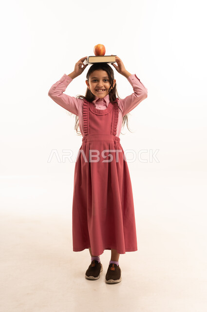 تناول الفواكه الصحية, الاستعداد للعام الدراسي الجديد، حب التعلم و التعليم, النظر إلى الكاميرا بإيماءات السعادة و السرور, بورتريه لفتاة عربية خليجية سعودية ترتدي الزي المدرسي تضع على رأسها الكتب المدرسية وتفاحة خضراء, الجسم بالطول الكامل, خلفية بيضاء