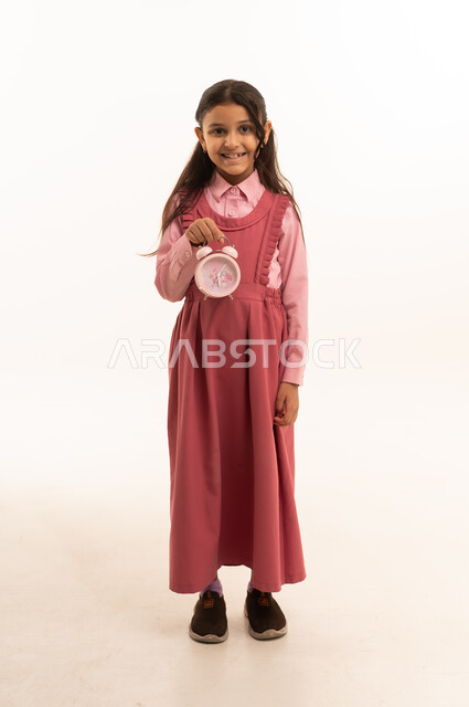 The concept of time management and organization, preparing for the new school year, education in the Kingdom of Saudi Arabia, portrait of a Saudi Arabian Gulf girl wearing a school uniform holding an alarm clock, full body, white background