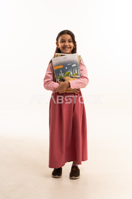 جودة التعليم في مدارس المملكة العربية السعودية، مفهوم الجد والاجتهاد للوصول إلى النجاح، العودة الى المدارس، بورتريه لطالبة عربيه خليجيه سعودية مبتسمه ترتدي الزي المدرسي تحمل مجموعة من الكتب، صورة لكامل الجسم، خلفية بيضاء