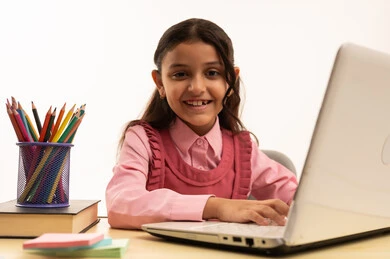 Use of modern technology, concept of distance learning, following online classes via the internet, portrait of a Saudi Arabian Gulf Arab female student wearing school uniform sitting at a study table working on a laptop with an expression of pleasure, white background