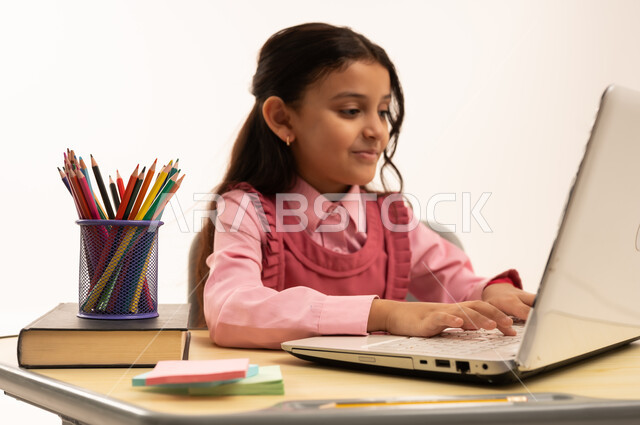 استخدام التكنولوجيا الحديثة، مفهوم التعلم عن بعد, متابعة حصص الاونلاين عن طريق الانترنت، بورتريه لطالبة عربية خليجية سعودية ترتدي اليونيفووم المدرسي تجلس على طاولة الدراسة   تعمل على جهاز الحاسوب المحمول بتعابير السرور، خلفية بيضاء