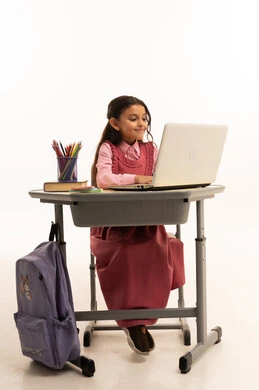 Use of modern technology, concept of distance learning, following online classes via the internet, portrait of a Saudi Arabian Gulf Arab female student wearing school uniform sitting at a study table working on a laptop with an expression of pleasure, white background