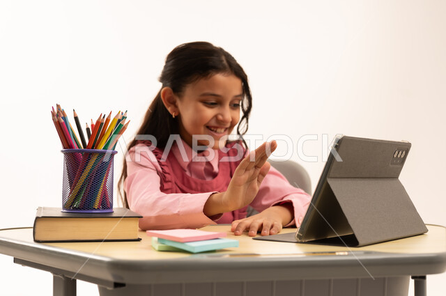 متابعة حصص الاونلاين عن طريق الانترنت, استخدام التكنولوجيا الحديثة، مفهوم التعلم عن بعد، بورتريه لطالبة عربية خليجية سعودية ترتدي اليونيفووم المدرسي تجلس على طاولة الدراسة   تعمل على جهاز التابلت اللوحي بتعابير السرور، خلفية بيضاء